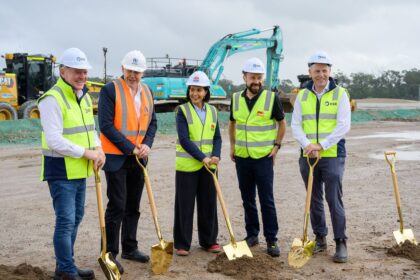 Five people wearing hard hats and high-vis vests hold ceremonial shovels at a construction site.