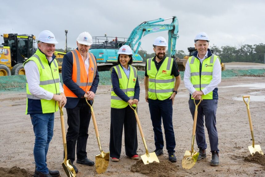 Five people wearing hard hats and high-vis vests hold ceremonial shovels at a construction site.