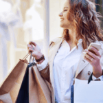 A smiling woman holding shopping bags and a smartphone outdoors.