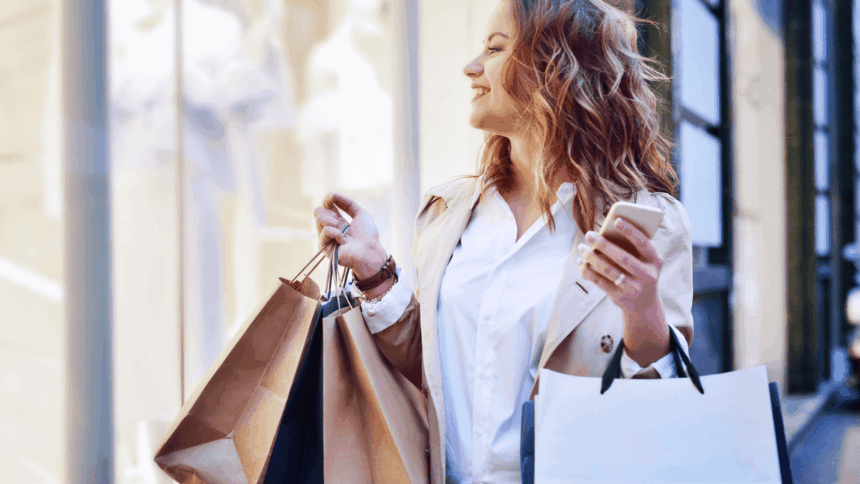 A smiling woman holding shopping bags and a smartphone outdoors.