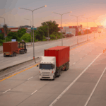 Semi-trucks hauling shipping containers travel on an open highway during a sunset.
