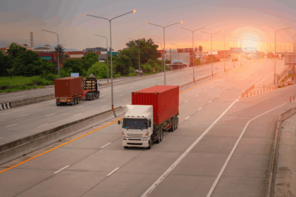 Semi-trucks hauling shipping containers travel on an open highway during a sunset.