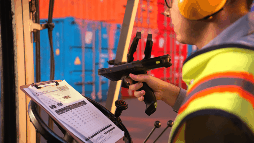 A worker in a high-visibility vest scans a shipping document at a container terminal.