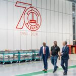 Three people walk through a warehouse past a large 70th-anniversary Al Sayer company logo.