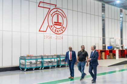 Three people walk through a warehouse past a large 70th-anniversary Al Sayer company logo.