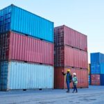 Two logistics professionals in hard hats walk among stacks of large shipping containers.