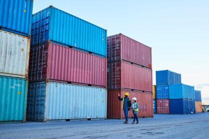 Two logistics professionals in hard hats walk among stacks of large shipping containers.