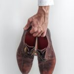 A hand in a white shirt holding a pair of brown leather dress shoes.