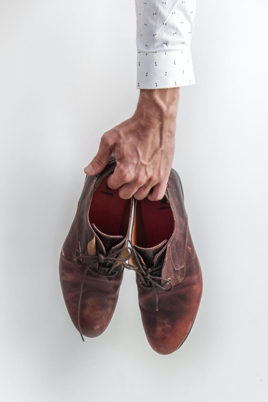 A hand in a white shirt holding a pair of brown leather dress shoes.