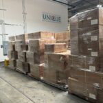 Stacked cardboard boxes wrapped in plastic inside a warehouse with a Unique sign.