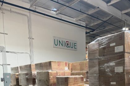 Stacked cardboard boxes wrapped in plastic inside a warehouse with a Unique sign.