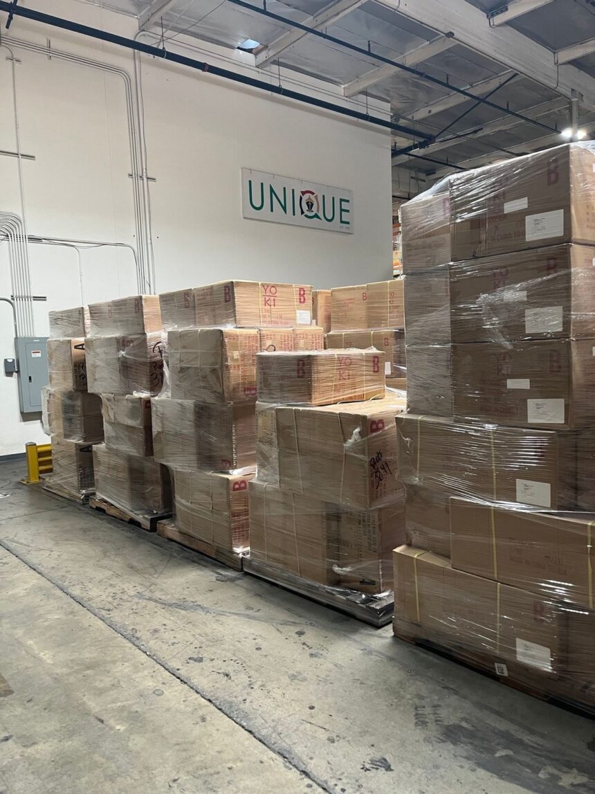 Stacked cardboard boxes wrapped in plastic inside a warehouse with a Unique sign.