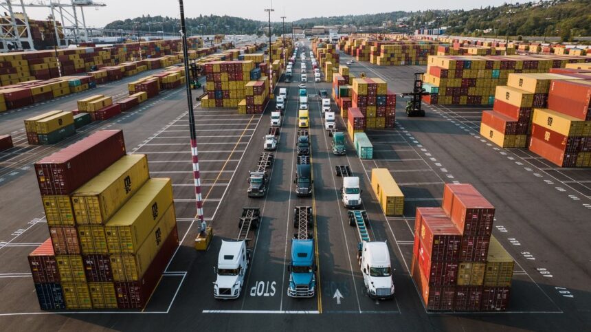Aerial view of semi-trucks queuing between rows of stacked shipping containers at a port.