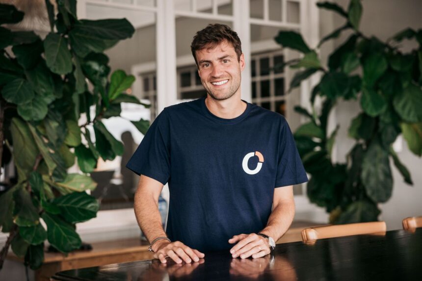 A smiling man wearing a dark blue t-shirt stands behind a dark table.