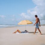 A woman relaxes under a beach umbrella while a man walks by on sand.