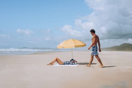 A woman relaxes under a beach umbrella while a man walks by on sand.