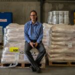 A man in a blue shirt sits on stacked pallets of wrapped goods.