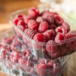 Two plastic containers stacked, filled with fresh red raspberries on a wooden surface.