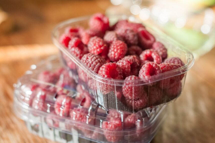 Two plastic containers stacked, filled with fresh red raspberries on a wooden surface.
