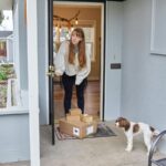 A woman opening her front door to several packages with a dog standing nearby.