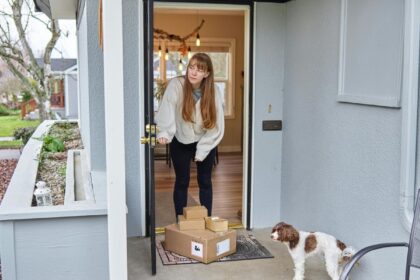 A woman opening her front door to several packages with a dog standing nearby.