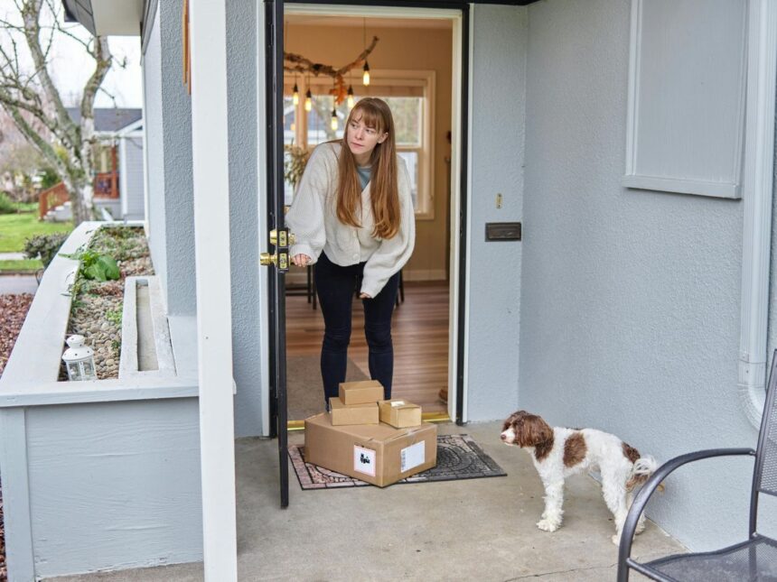 A woman opening her front door to several packages with a dog standing nearby.