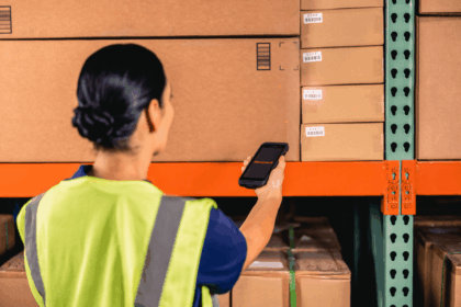 A warehouse worker in a high-visibility vest scans a cardboard box with a scanner.