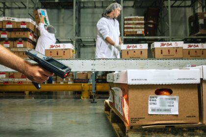 Hand holding a barcode scanner aimed at crates of produce in a warehouse.