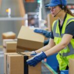 A warehouse worker in a high-visibility vest placing boxes onto a conveyor belt.