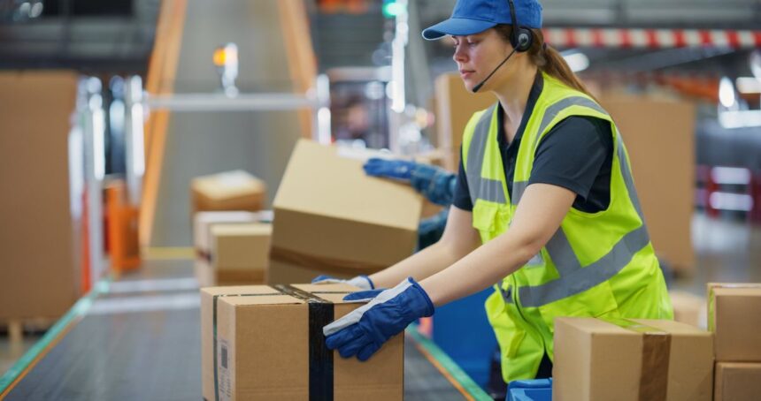 A warehouse worker in a high-visibility vest placing boxes onto a conveyor belt.