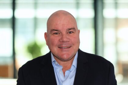 A smiling, bald man wearing a light blue dress shirt and navy blue suit jacket.
