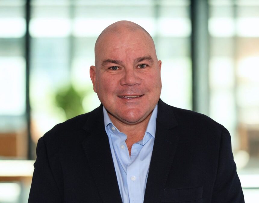 A smiling, bald man wearing a light blue dress shirt and navy blue suit jacket.