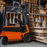 Orange forklift in a warehouse with stacked wooden panels and a blurred worker.