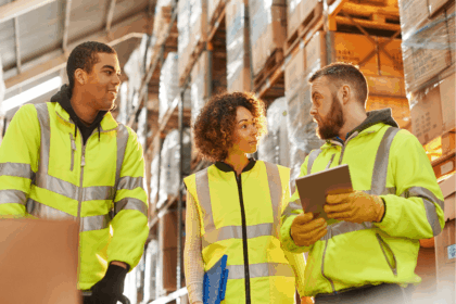 Three warehouse workers in high-visibility vests discussing logistics in a storage facility.