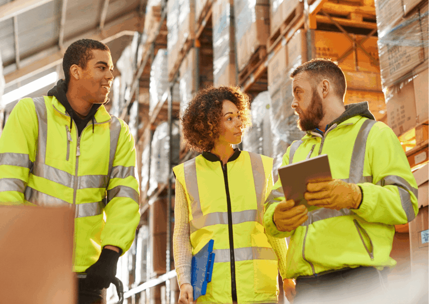 Three warehouse workers in high-visibility vests discussing logistics in a storage facility.