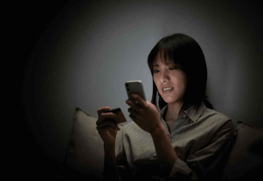 A woman shops online using her smartphone and a credit card in the dark.
