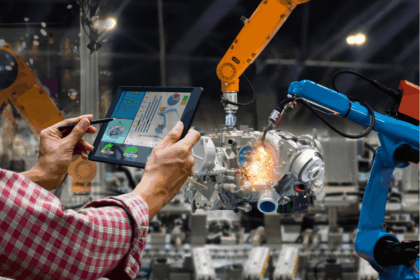 A person uses a tablet to monitor industrial robotic arms welding a car engine.