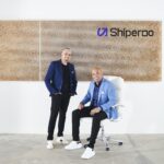 Two men in business casual attire pose in front of a Shiperoo office logo wall.