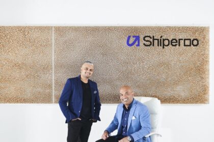 Two men in business casual attire pose in front of a Shiperoo office logo wall.
