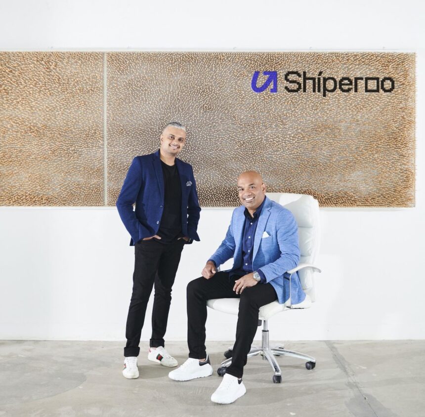 Two men in business casual attire pose in front of a Shiperoo office logo wall.