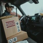 Delivery driver in a van with stacked cardboard packages, one marked Fragile.