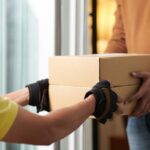 A delivery worker in gloves handing a cardboard box to a customer.