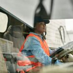 A woman wearing a cap and high-visibility vest driving a truck.