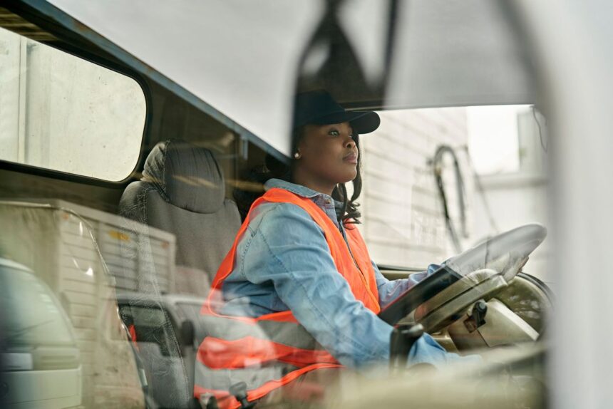 A woman wearing a cap and high-visibility vest driving a truck.