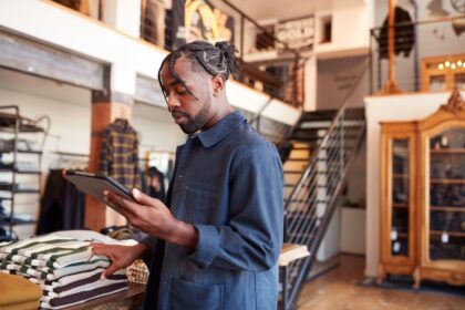 A man wearing a blue jacket uses a tablet while organizing sweaters in a store.