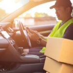 Delivery driver in a vehicle checking a smartphone with packages on the seat.