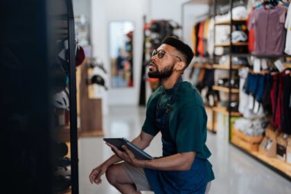 A shop worker holding a tablet checks merchandise on a shelf in a store.