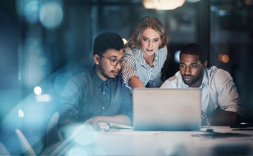 Three diverse colleagues collaborating on a project on a laptop in a dim office.