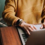 Person wearing a mustard sweater and gold bracelets typing on a laptop.