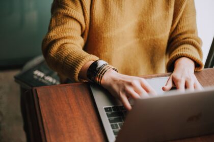 Person wearing a mustard sweater and gold bracelets typing on a laptop.